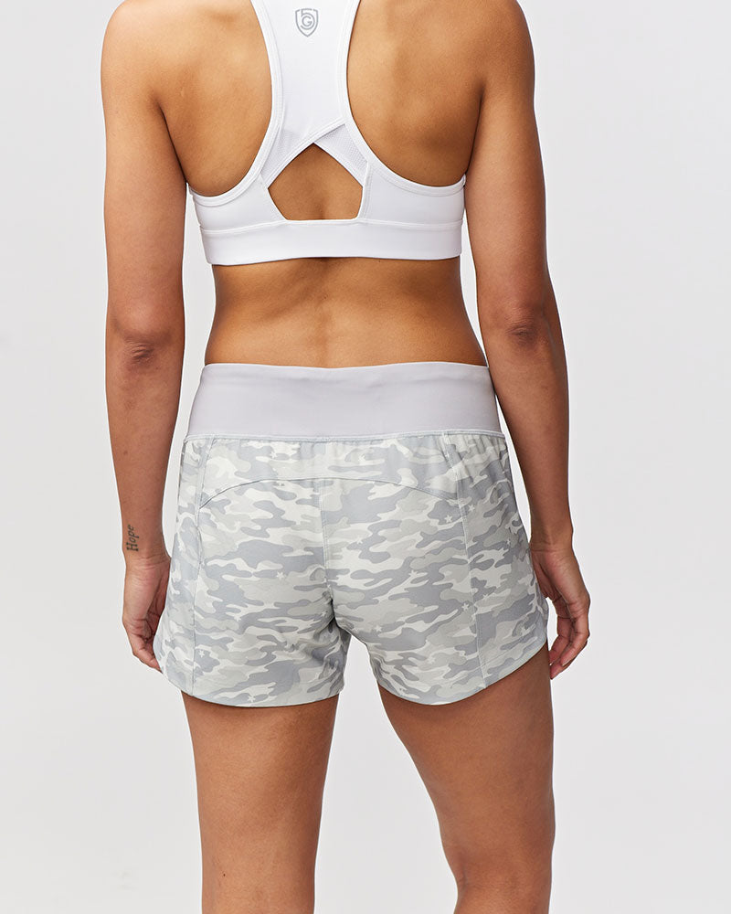 Back view of a woman wearing Excel Sculpt Performance Shorts with a light camouflage pattern and a white sports bra.
